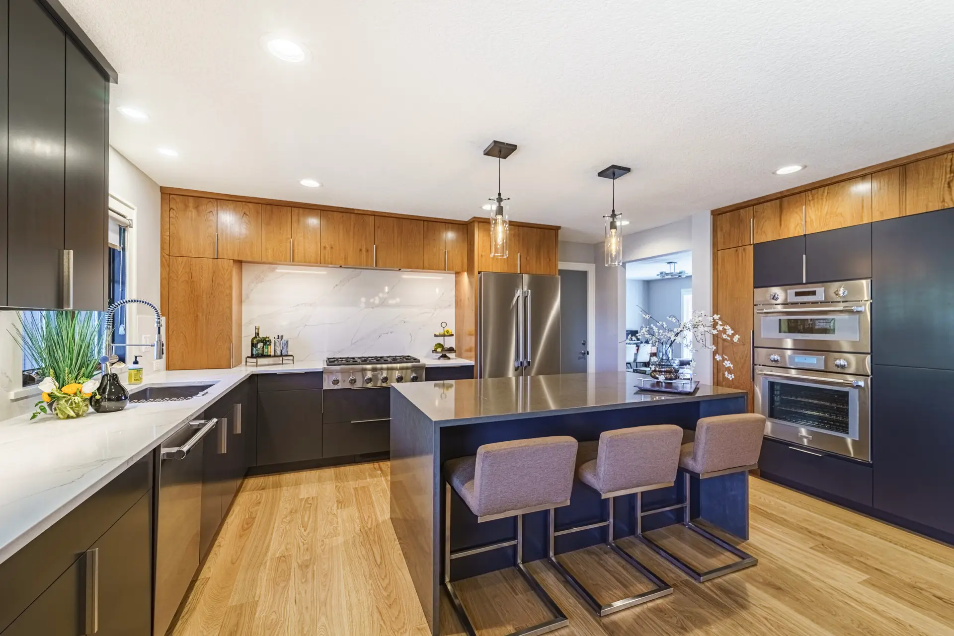 Modern kitchen with black lower cabinets, wooden upper cabinets, and a center island with a steel countertop. A stainless oven and fridge line the right wall; pendant lights hang from the ceiling.