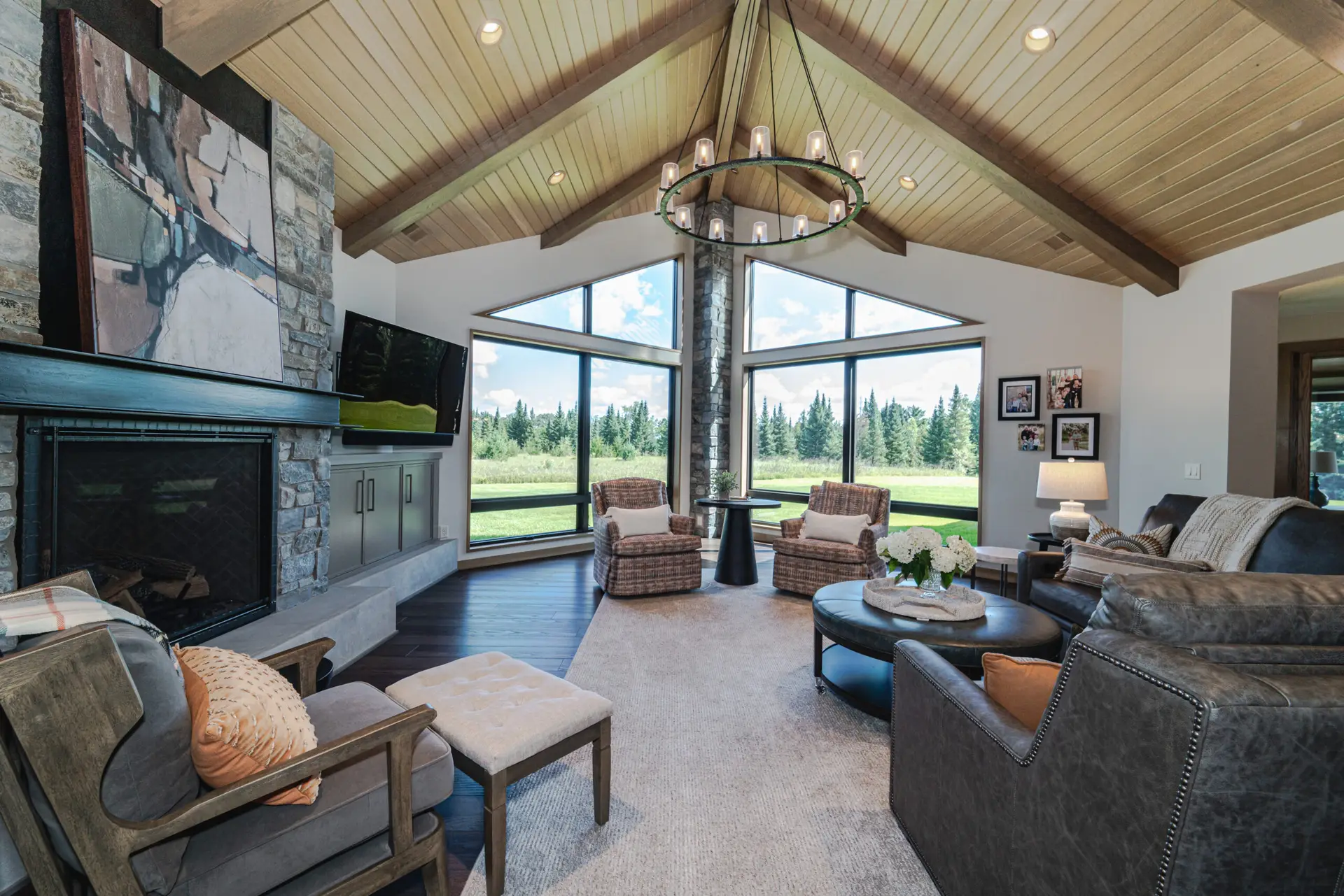 Bright living room with a stone fireplace, large triangular windows, and a circular chandelier overhead; outdoors shows trees and blue sky.