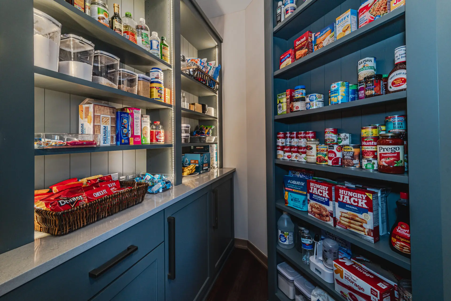 Blue kitchen pantry with tall shelves stocked with cans, boxes, jars, and snack bags along both walls.
