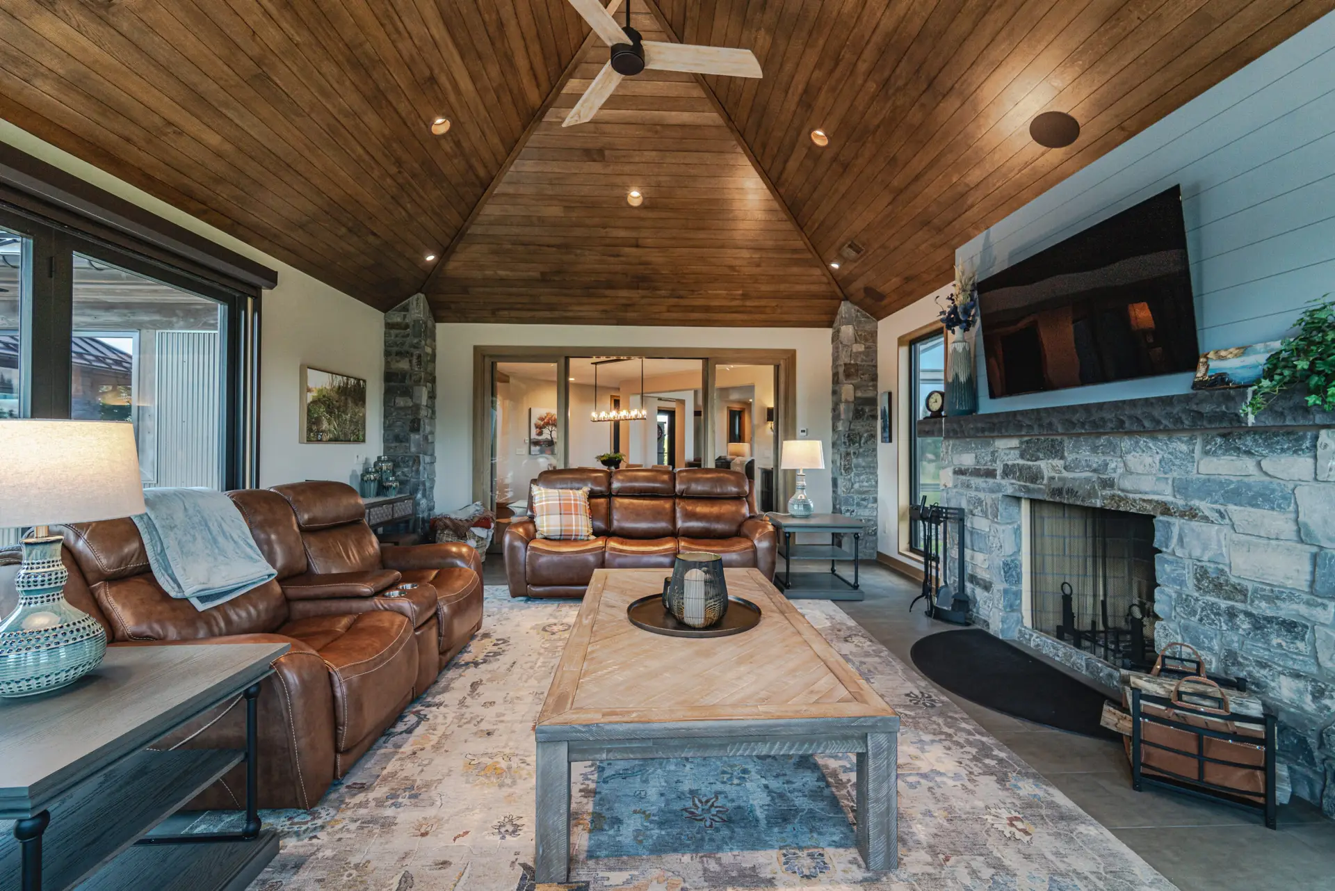 Cozy living room with a vaulted wooden ceiling, stone fireplace, and brown leather seating arrangement.
