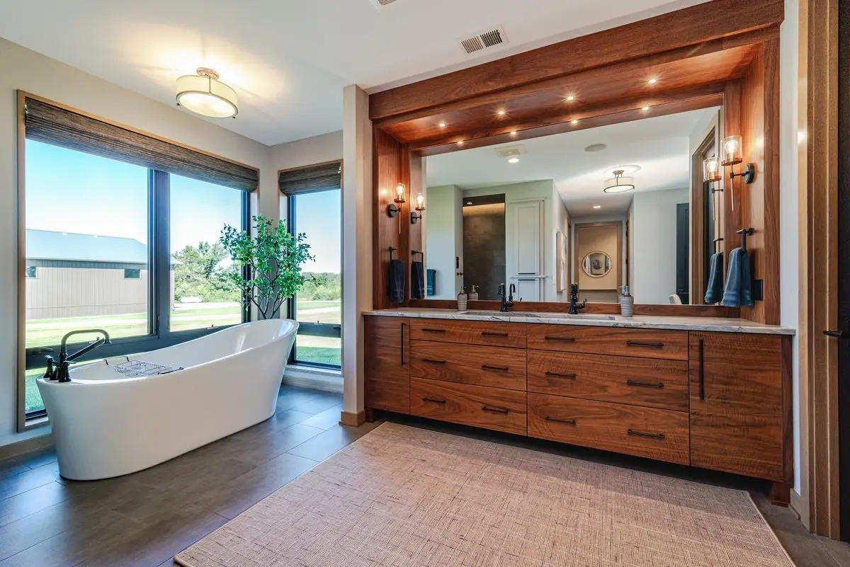 Bright bathroom with a white freestanding tub by large windows and a long wood vanity with dual sinks and a wide mirror callibrationed by warm lighting.