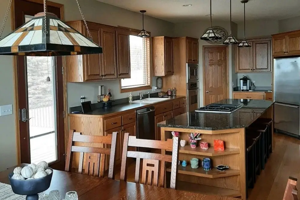 Bright kitchen with wooden cabinets and a central granite island, hanging pendant lights, and a wooden dining table nearby.