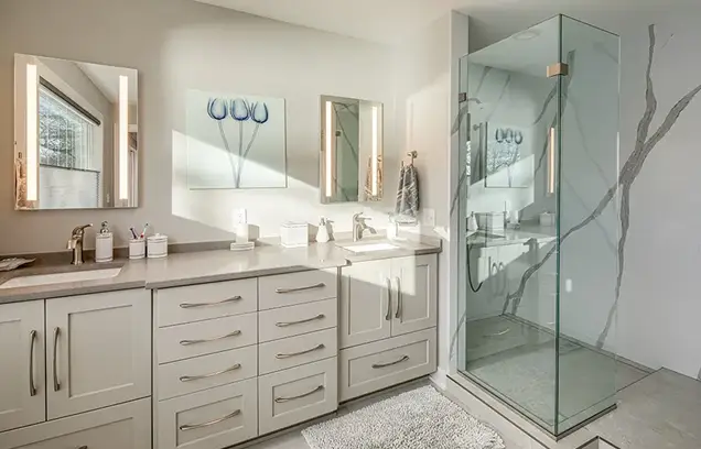 Bright modern bathroom with a double-sink vanity, white cabinets, backlit mirrors, marble countertop, and a glass-walled walk-in shower.