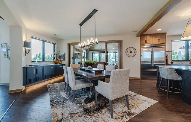 Open-concept dining area: dark wood table with eight beige chairs on a patterned rug, chandelier, and a kitchen with dark cabinets and stainless appliances.
