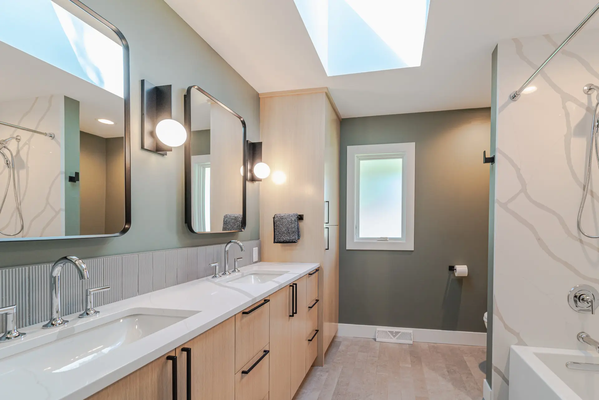 Modern bathroom with a double sink vanity, light wood cabinets, two mirrors, globe lights, gray walls, a skylight, and a glass shower on the right.