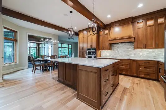 Kitchen with a large island and wood cabinets, pendant lights, and a dining area visible in the background; light hardwood floors throughout.