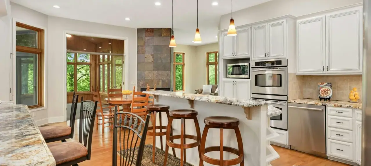 Bright modern kitchen with white cabinets, granite island, and stainless steel appliances; wooden bar stools along the island.