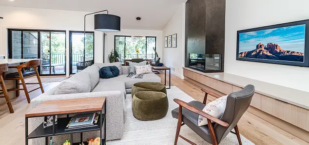 Open-plan living room: gray sectional sofa, olive ottomans, wooden lounge chair, fireplace with built-in storage, wall-mounted TV, and large windows/sliding doors to a balcony.