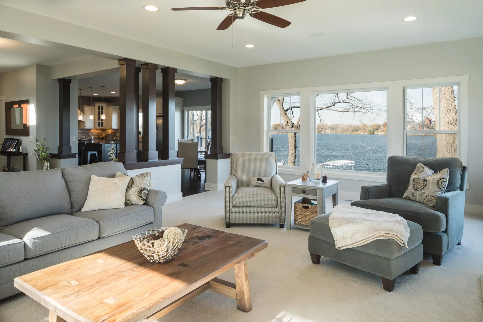 Open-concept living room with gray sofas, a wood coffee table, and a pair of patterned armchairs, overlooking large windows to a lake beyond.